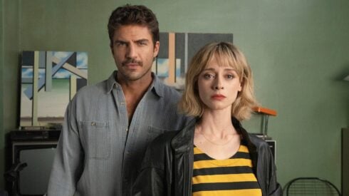 A manin a grey shirt and woman in a yellow and blue striped top pose for a promotional photo in front of a green wall.